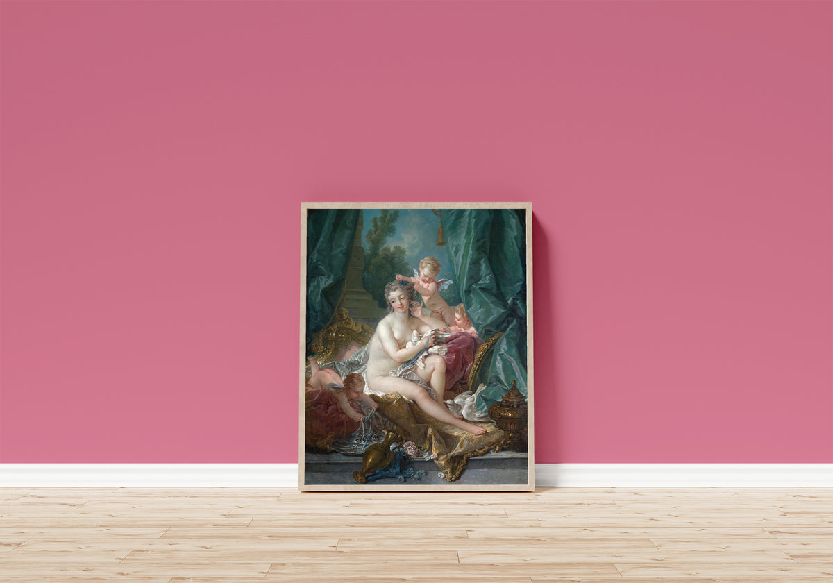 Framed art print of The Toilette of Venus by François Boucher, in a light wood frame leaning on a pink wall above a wooden floor