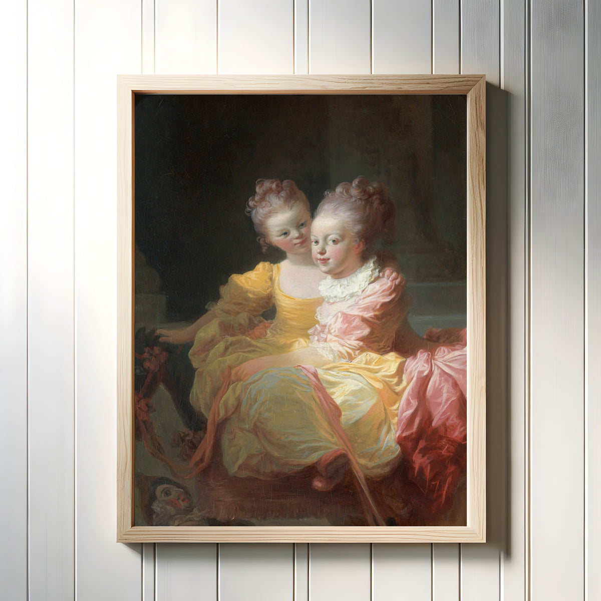 Framed art print of The Two Sisters by Jean-Honoré Fragonard, in a light wood frame on a white panelled wall