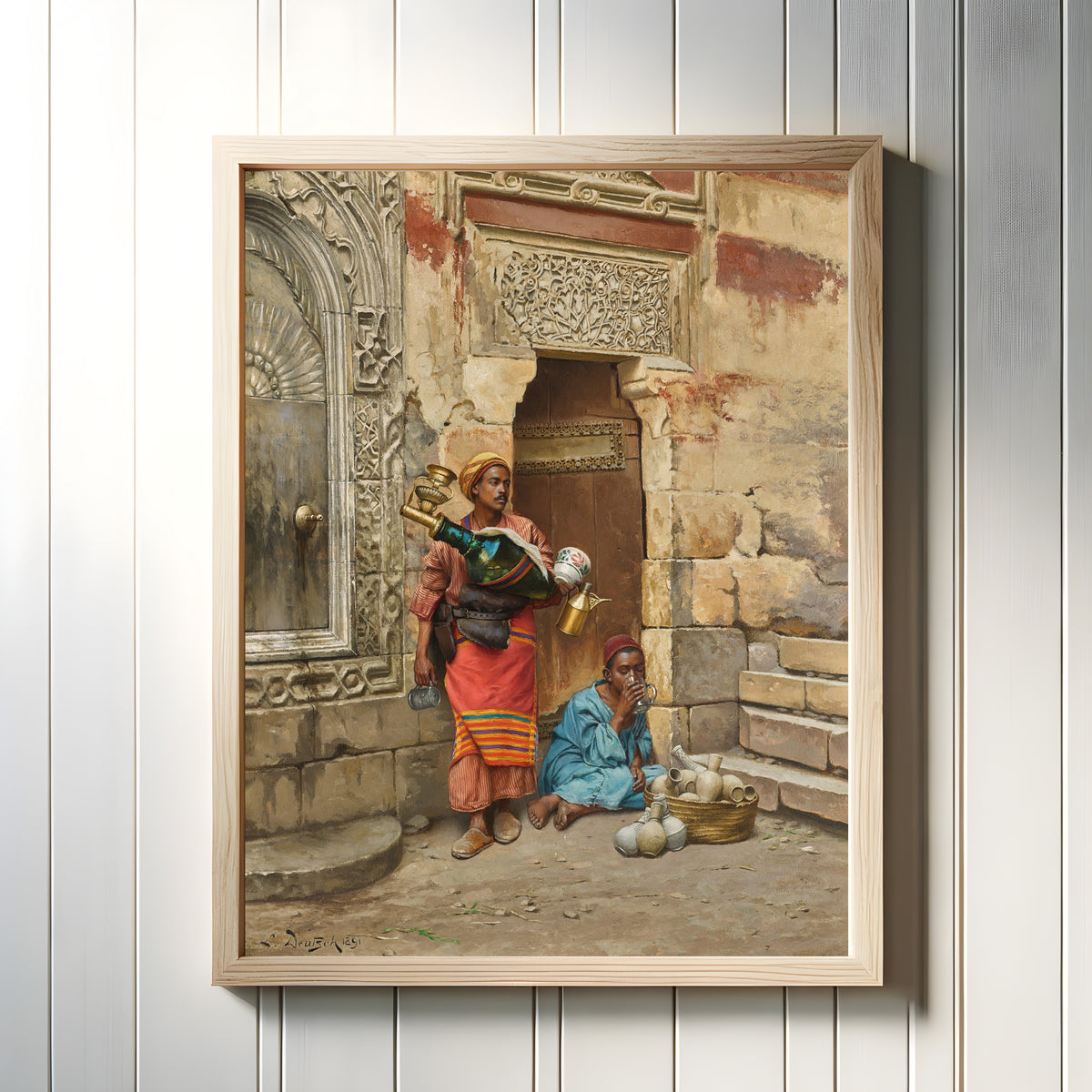 Framed art print of The Water Seller by Ludwig Deutsch, in a light wood frame on a white panelled wall