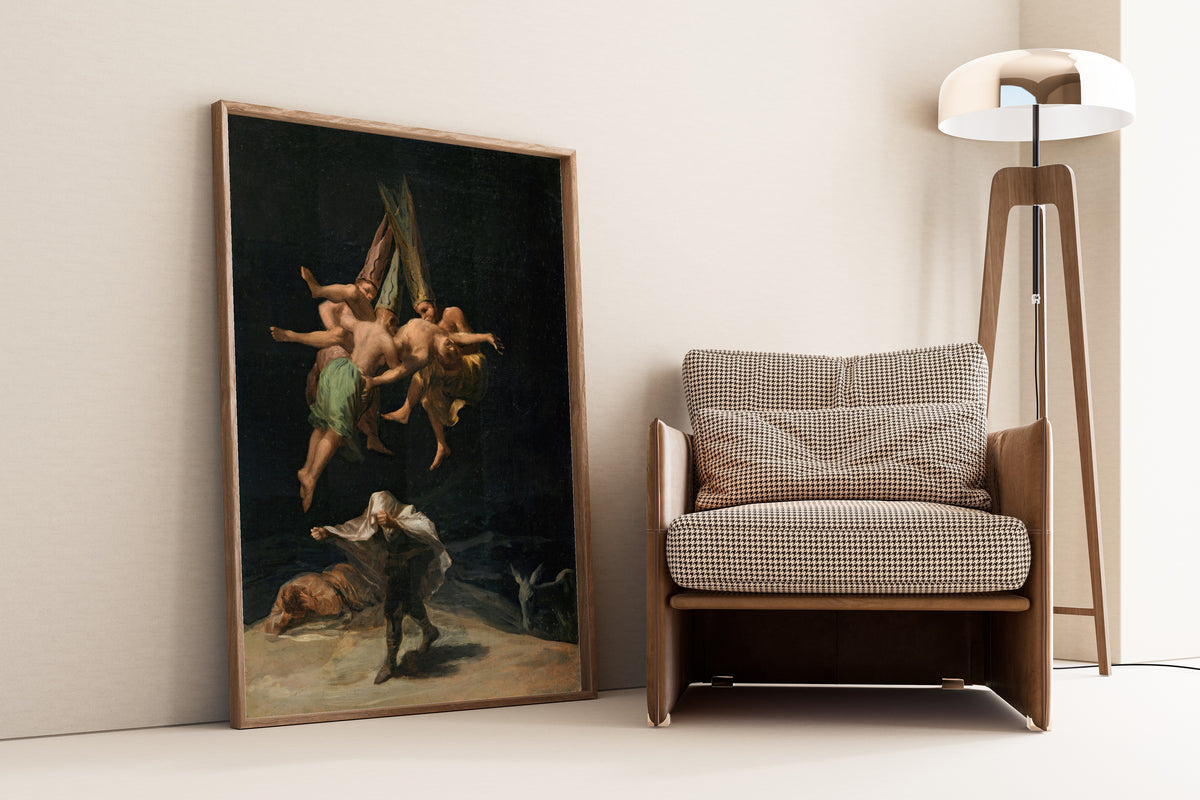 Framed art print of Witches' Flight by Francisco de Goya, leaning against a wall beside an armchair and a floor lamp