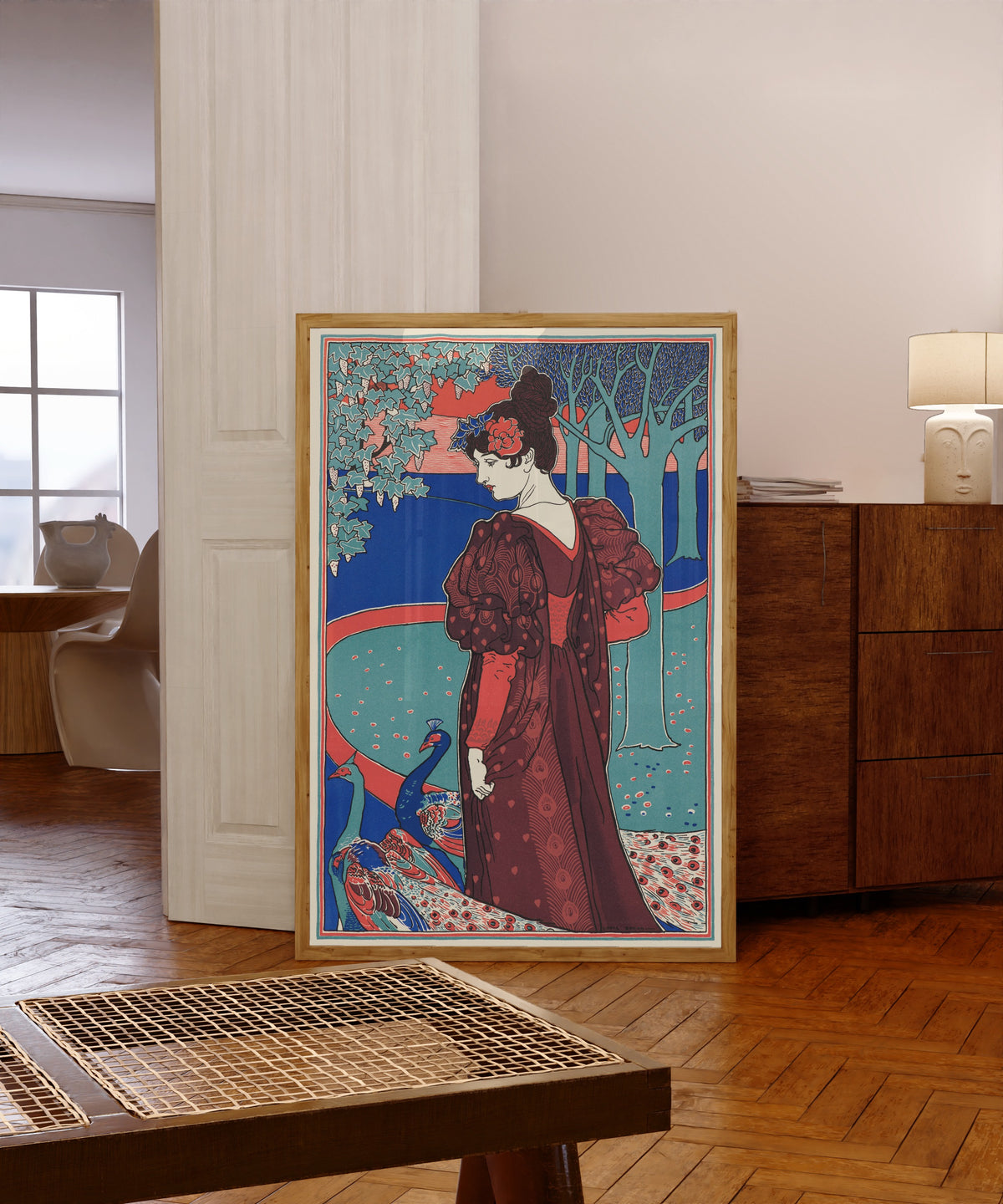 Framed art print of Woman with Peacocks (La Femme au Paon) by Louis Rhead, in a light wood frame leaning in a warm room with a parquet floor