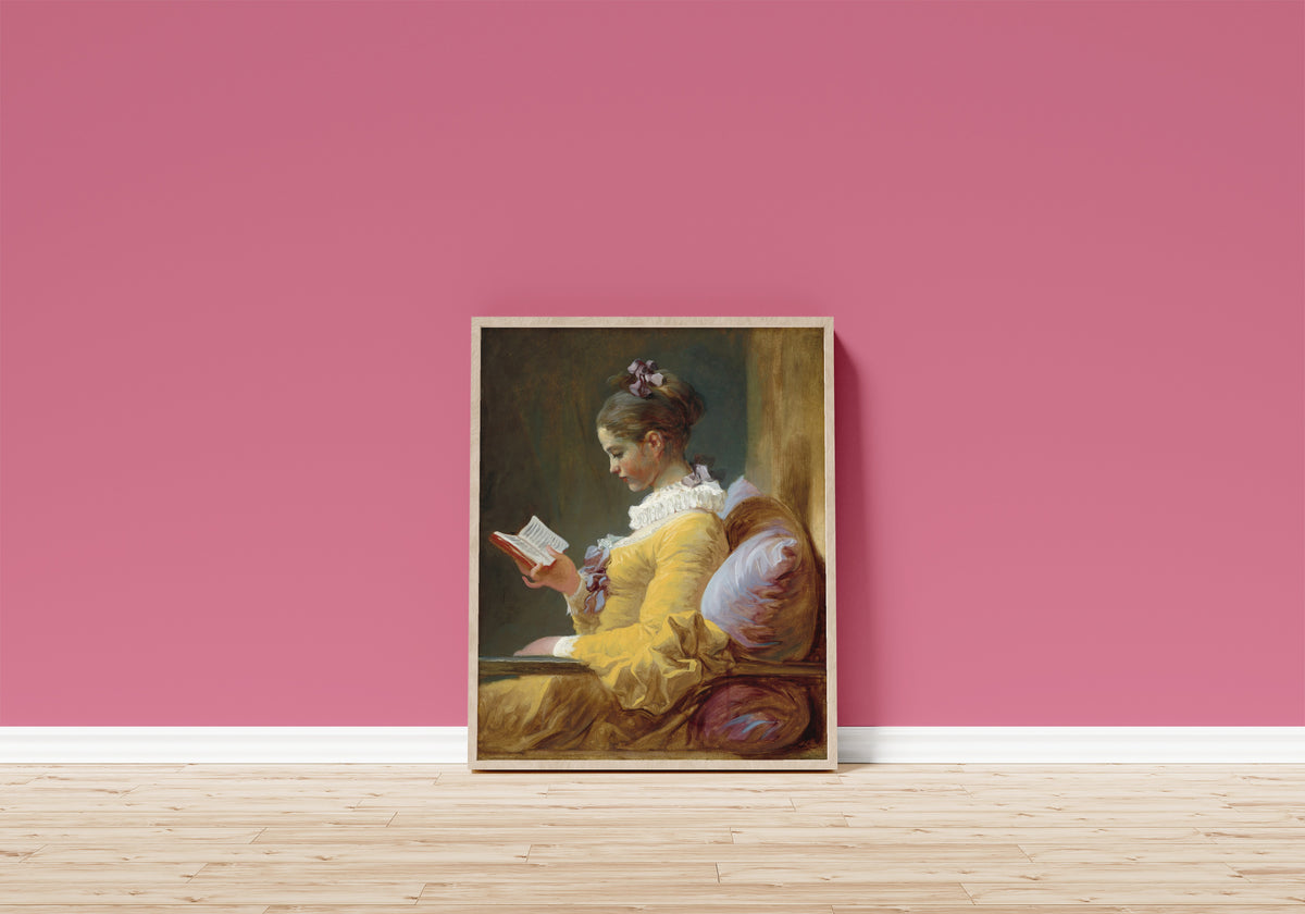 Framed art print of Young Girl Reading by Jean-Honoré Fragonard, in a light wood frame leaning on a pink wall above a wooden floor