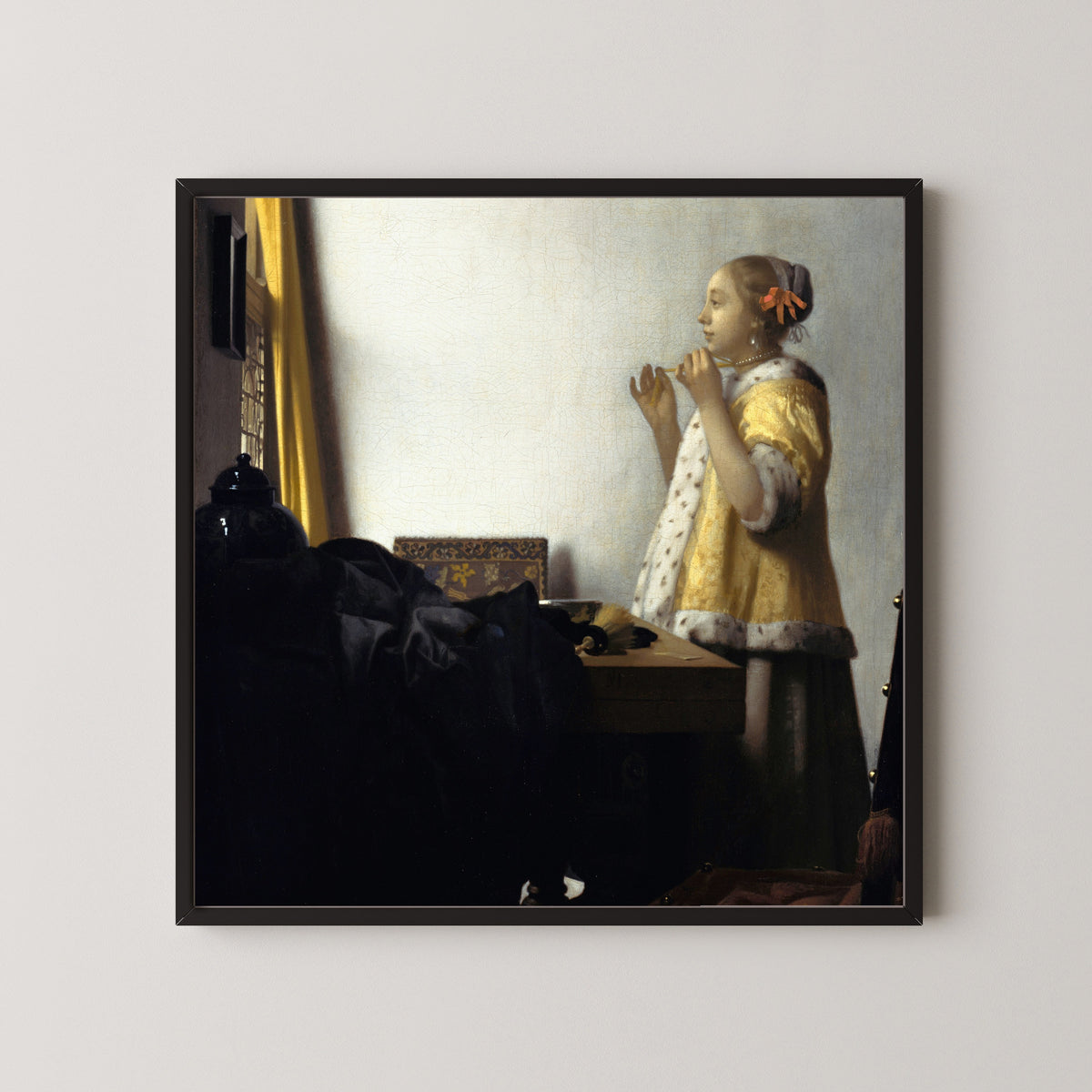 Framed art print of Young Woman with a Pearl Necklace by Johannes Vermeer, in a black frame on a pale wall