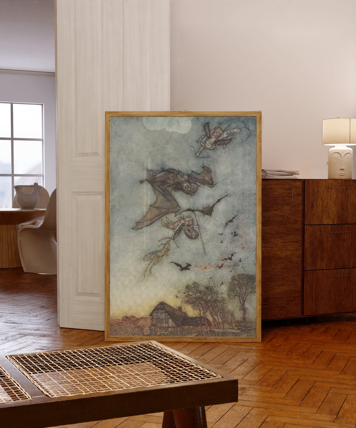 Framed art print of Some War with Rere-Mice for Their Leathern Wings by Arthur Rackham, in a light wood frame leaning in a warm room with a parquet floor