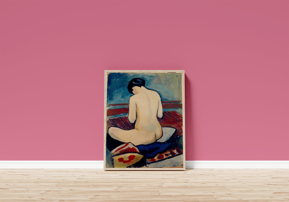 Framed art print of Sitting Nude with Pillow by August Macke, in a light wood frame leaning on a pink wall above a wooden floor