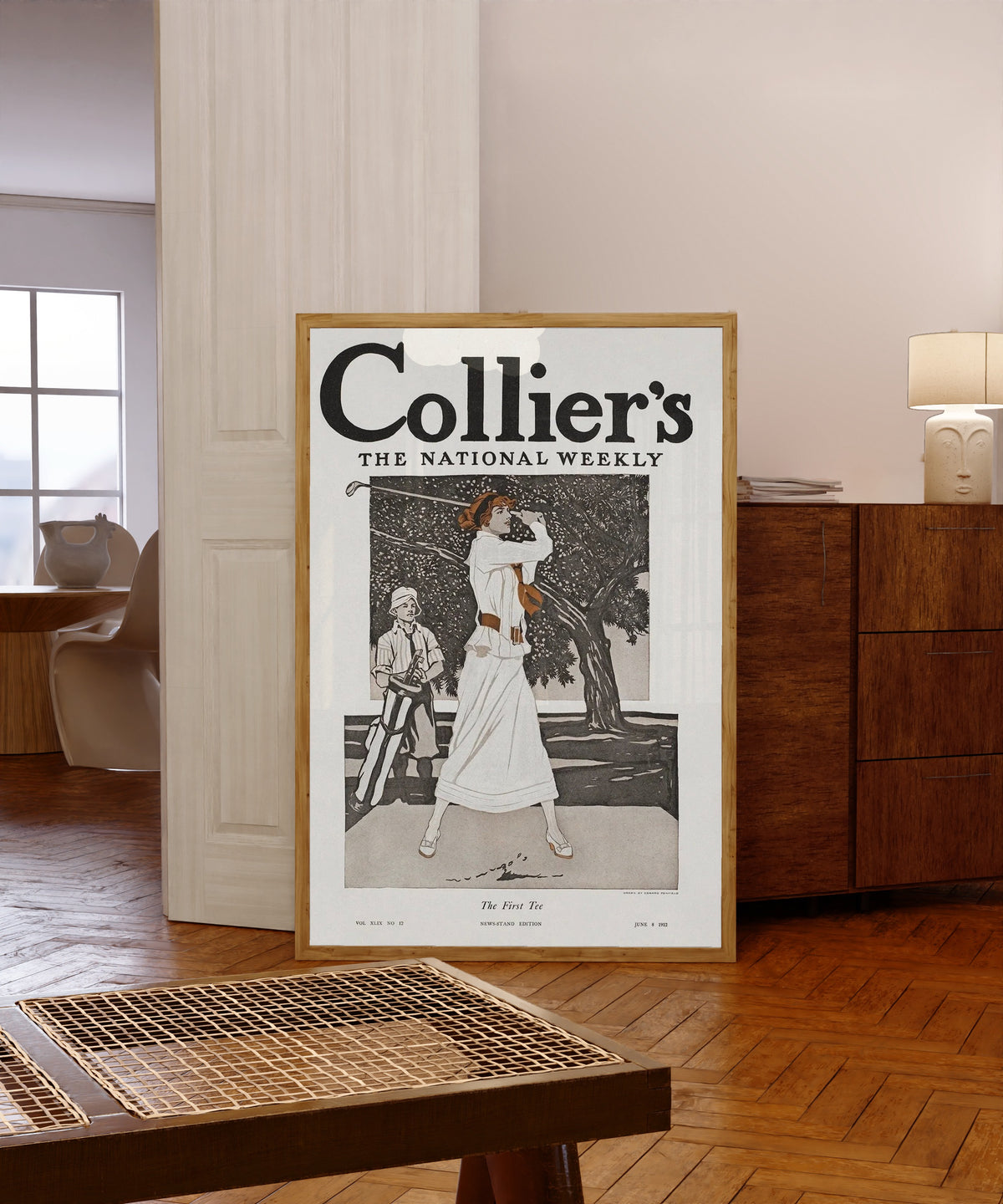 Framed art print of Lady Golfer (The First Tee) by Edward Penfield, in a light wood frame leaning in a warm room with a parquet floor