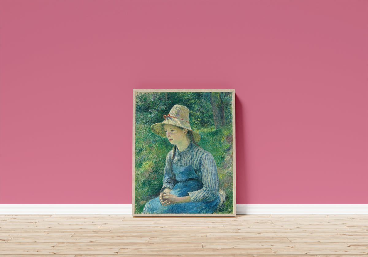 Framed art print of Peasant Girl with a Straw Hat by Camille Pissarro, in a light wood frame leaning on a pink wall above a wooden floor