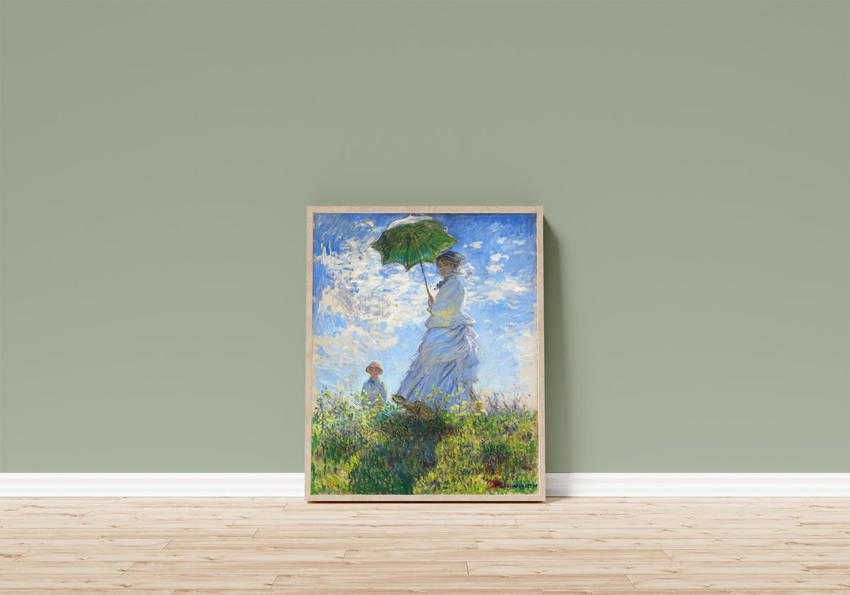 Framed art print of Woman with a Parasol - Madame Monet and Her Son by Claude Monet, in a light wood frame leaning on a sage green wall above a wooden floor