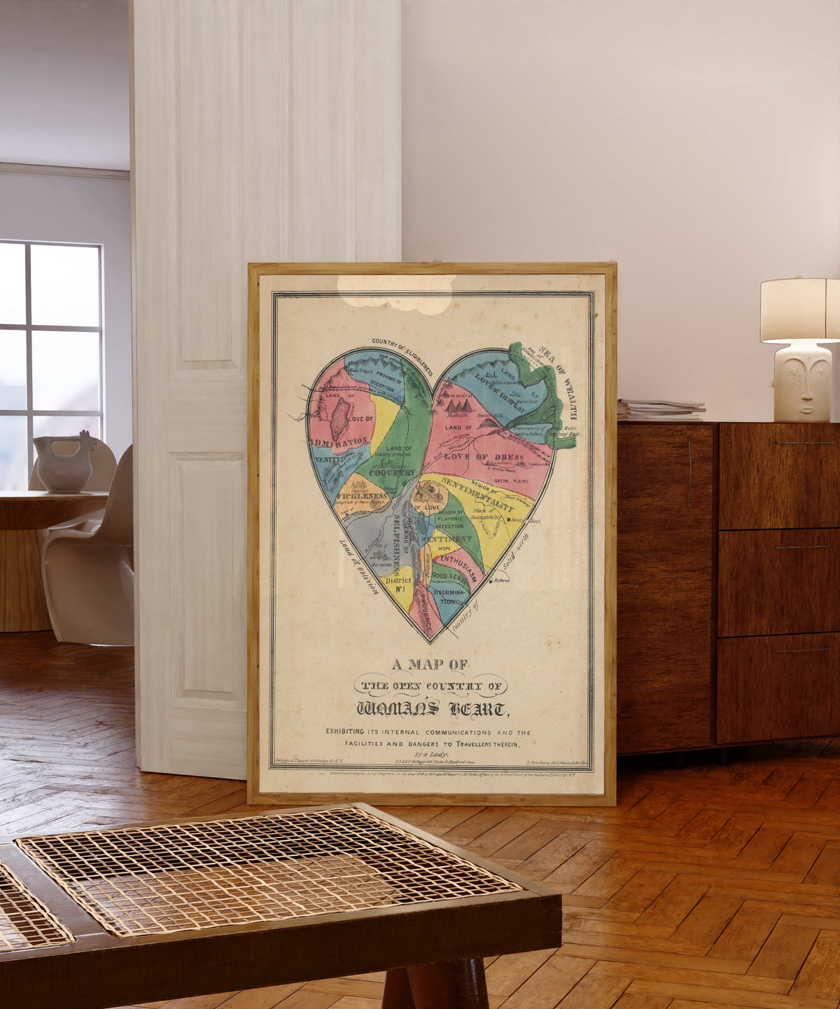 Framed art print of A Map of the Open Country of Woman's Heart by E.B. & E.C. Kellogg, in a light wood frame leaning in a warm room with a parquet floor