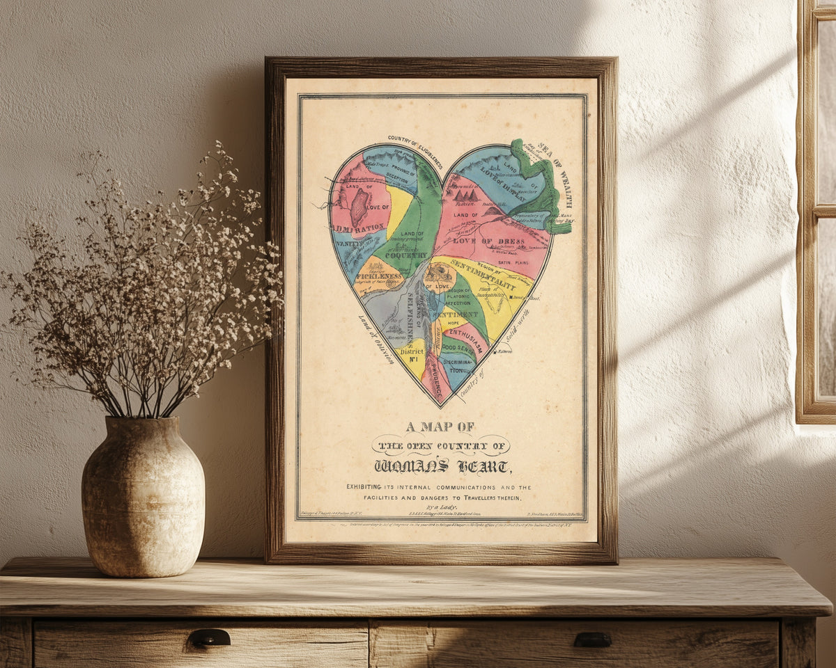 Framed art print of A Map of the Open Country of Woman's Heart by E.B. & E.C. Kellogg, in a dark wood frame leaning on a rustic console beside dried flowers