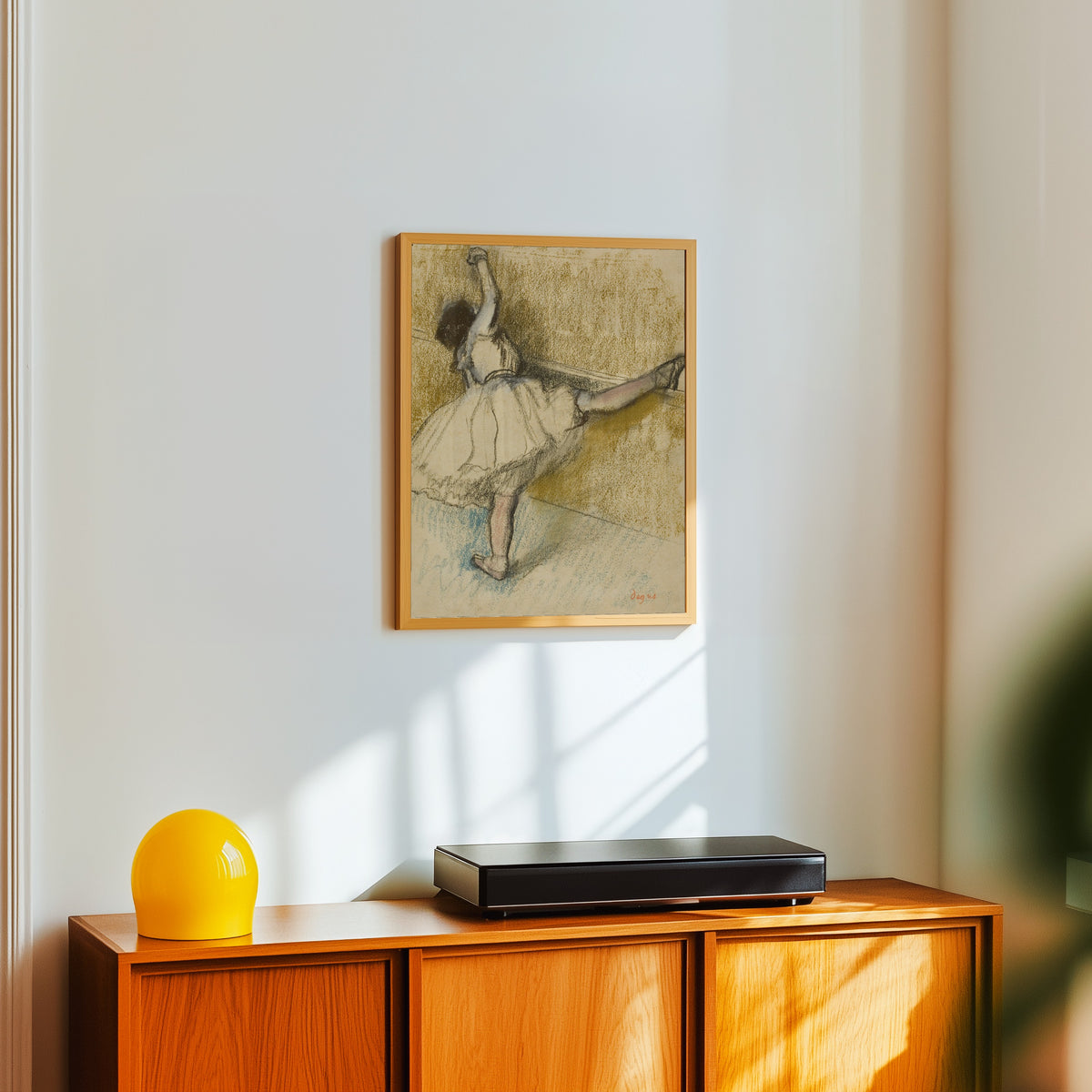 Framed art print of Dancer Stretching at the Bar by Edgar Degas, in a light wood frame on a white wall above an orange sideboard