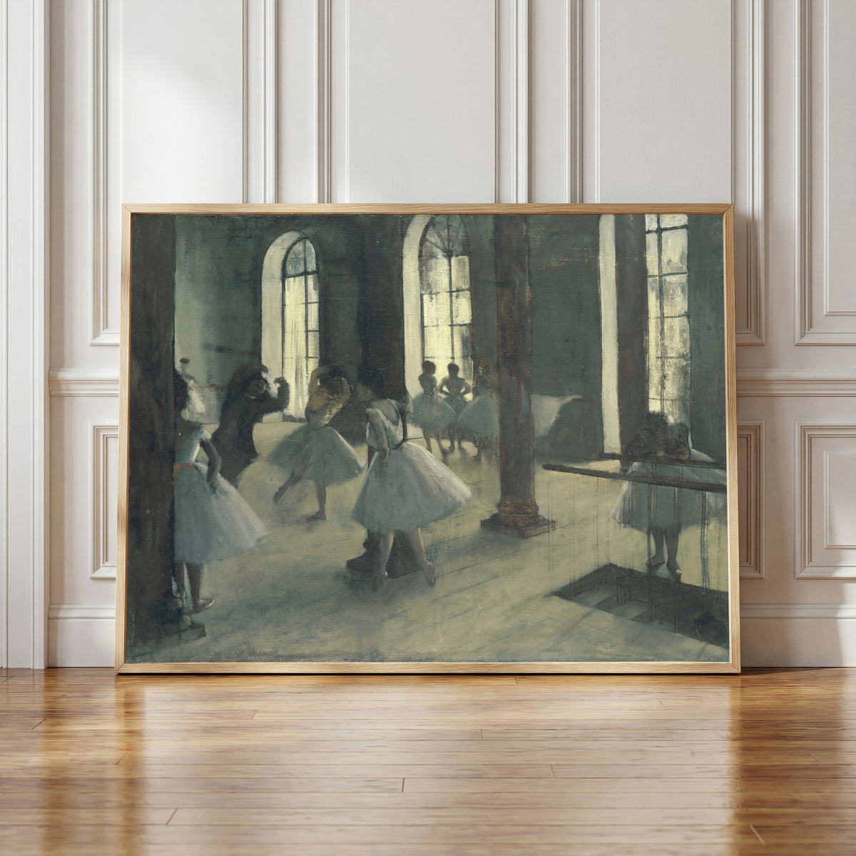 Framed art print of La Répétition au foyer de la danse by Edgar Degas, in a light wood frame leaning against a panelled white wall