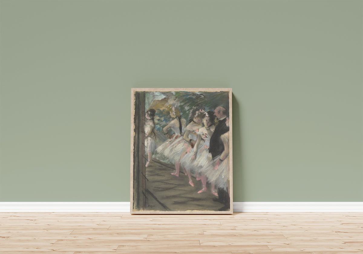 Framed art print of The Ballet by Edgar Degas, in a light wood frame leaning on a sage green wall above a wooden floor