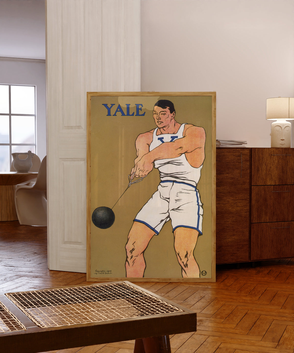 Framed art print of Yale by Edward Penfield, in a light wood frame leaning in a warm room with a parquet floor