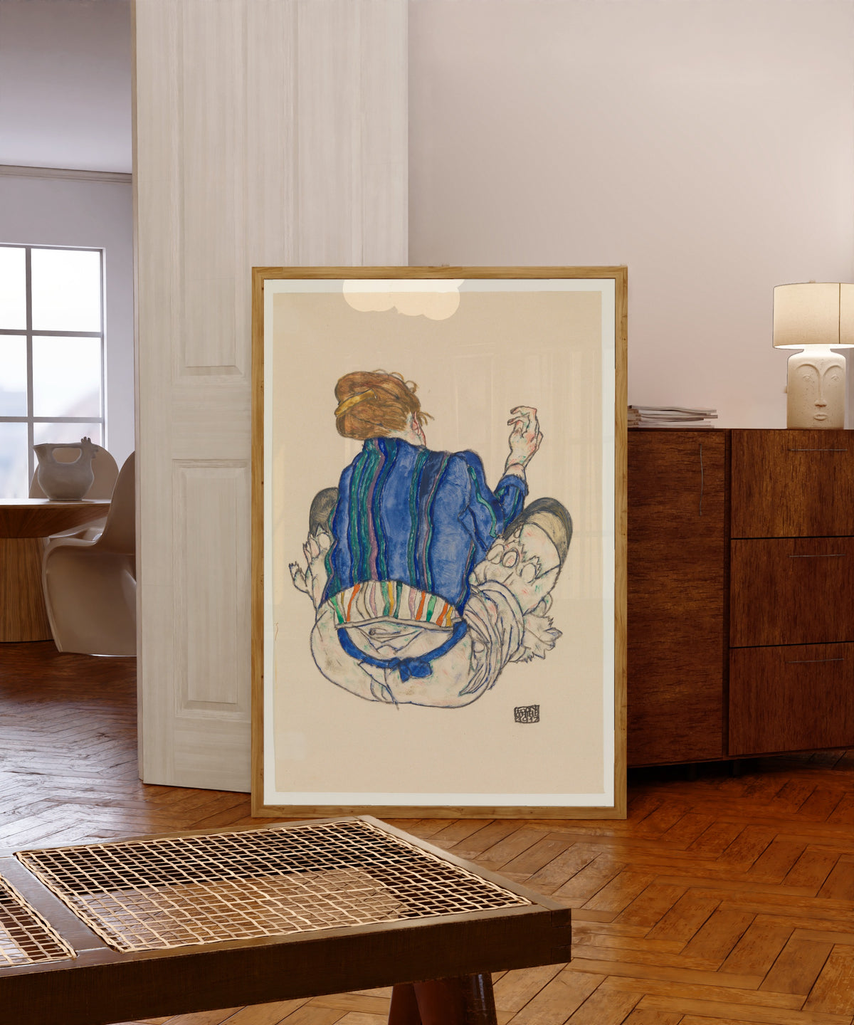 Framed art print of Seated Woman (blue jacket) by Egon Schiele, in a light wood frame leaning in a warm room with a parquet floor