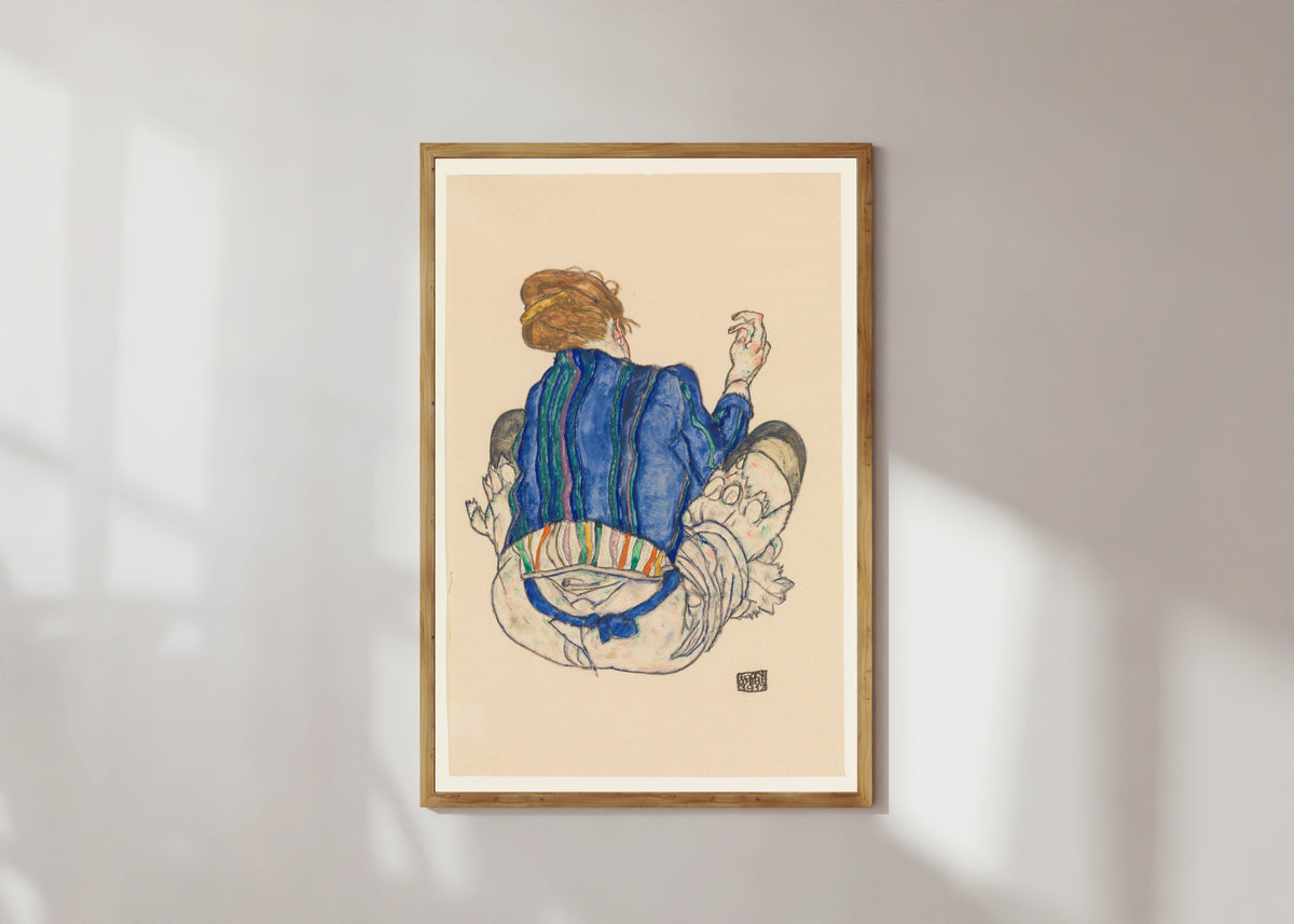 Framed art print of Seated Woman (blue jacket) by Egon Schiele, in a light wood frame on a sunlit white wall