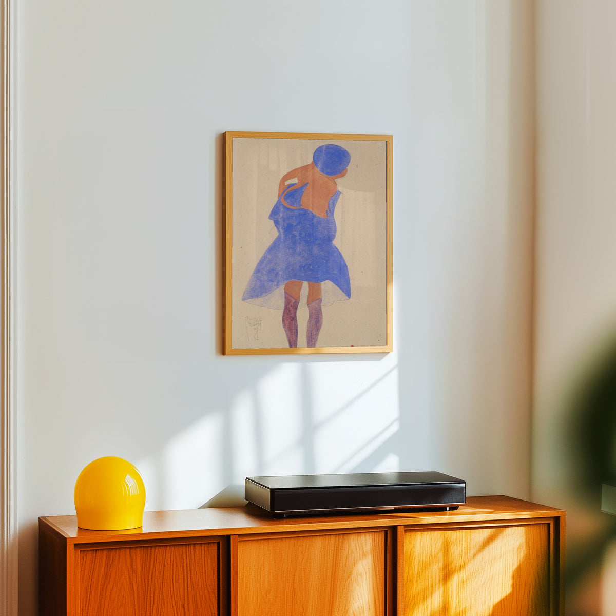 Framed art print of Standing Girl, Back View by Egon Schiele, in a light wood frame on a white wall above an orange sideboard