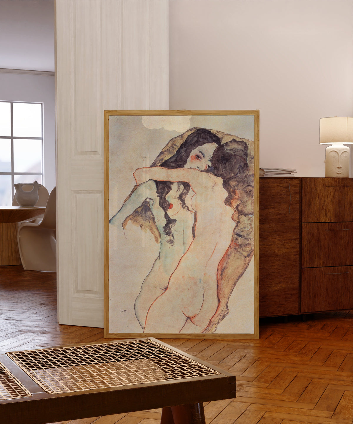 Framed art print of Two Embracing Women by Egon Schiele, in a light wood frame leaning in a warm room with a parquet floor