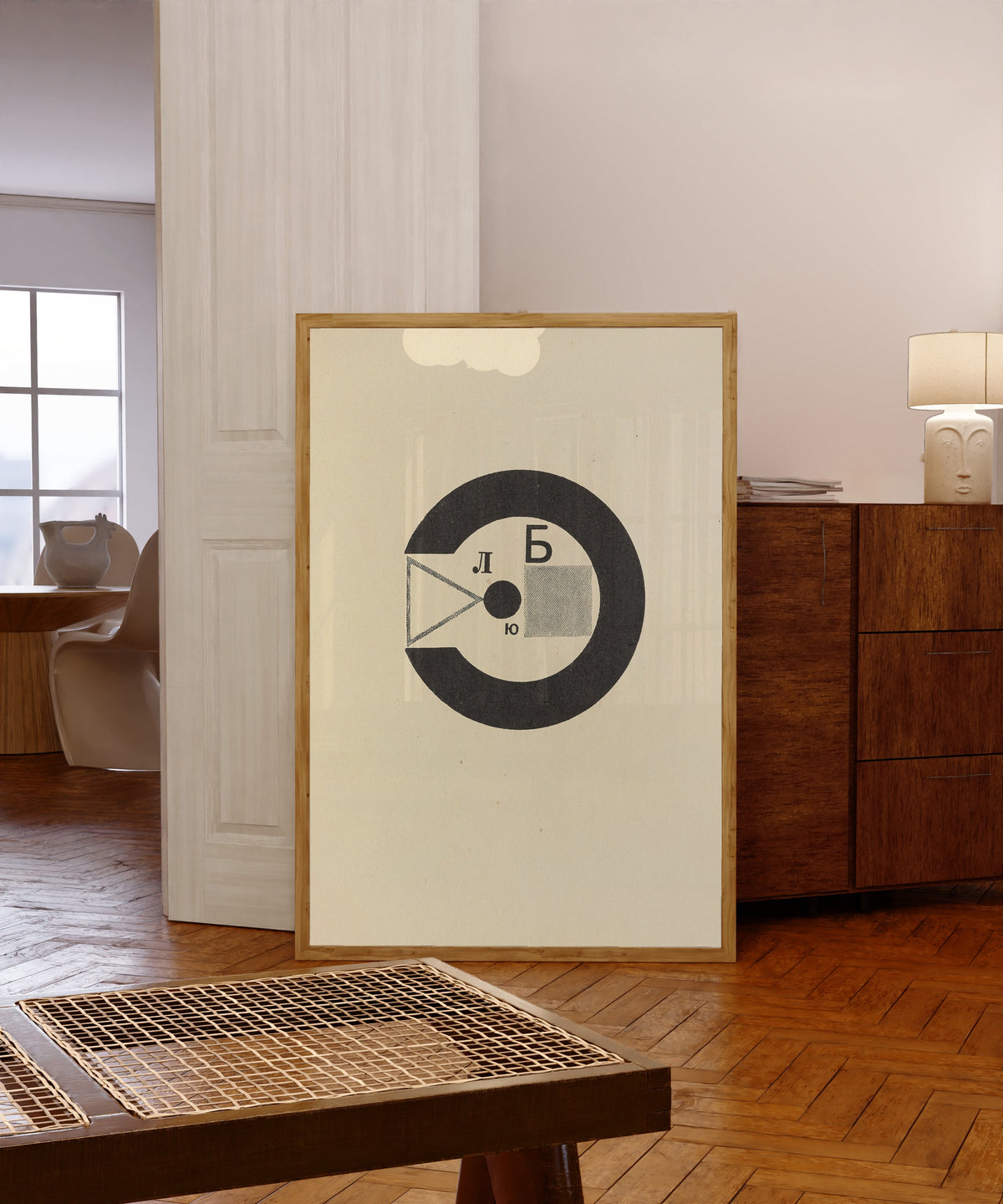 Framed art print of For the Voice: Black Ring with Letters by El Lissitzky, in a light wood frame leaning in a warm room with a parquet floor