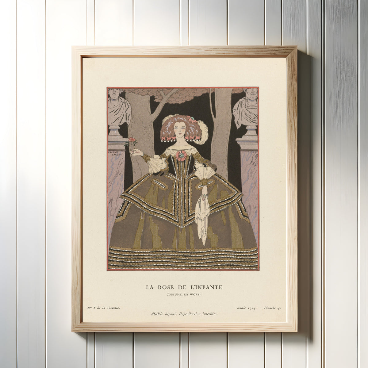 Framed art print of La Rose de l'Infante, costume de Worth by George Barbier, in a light wood frame on a white panelled wall