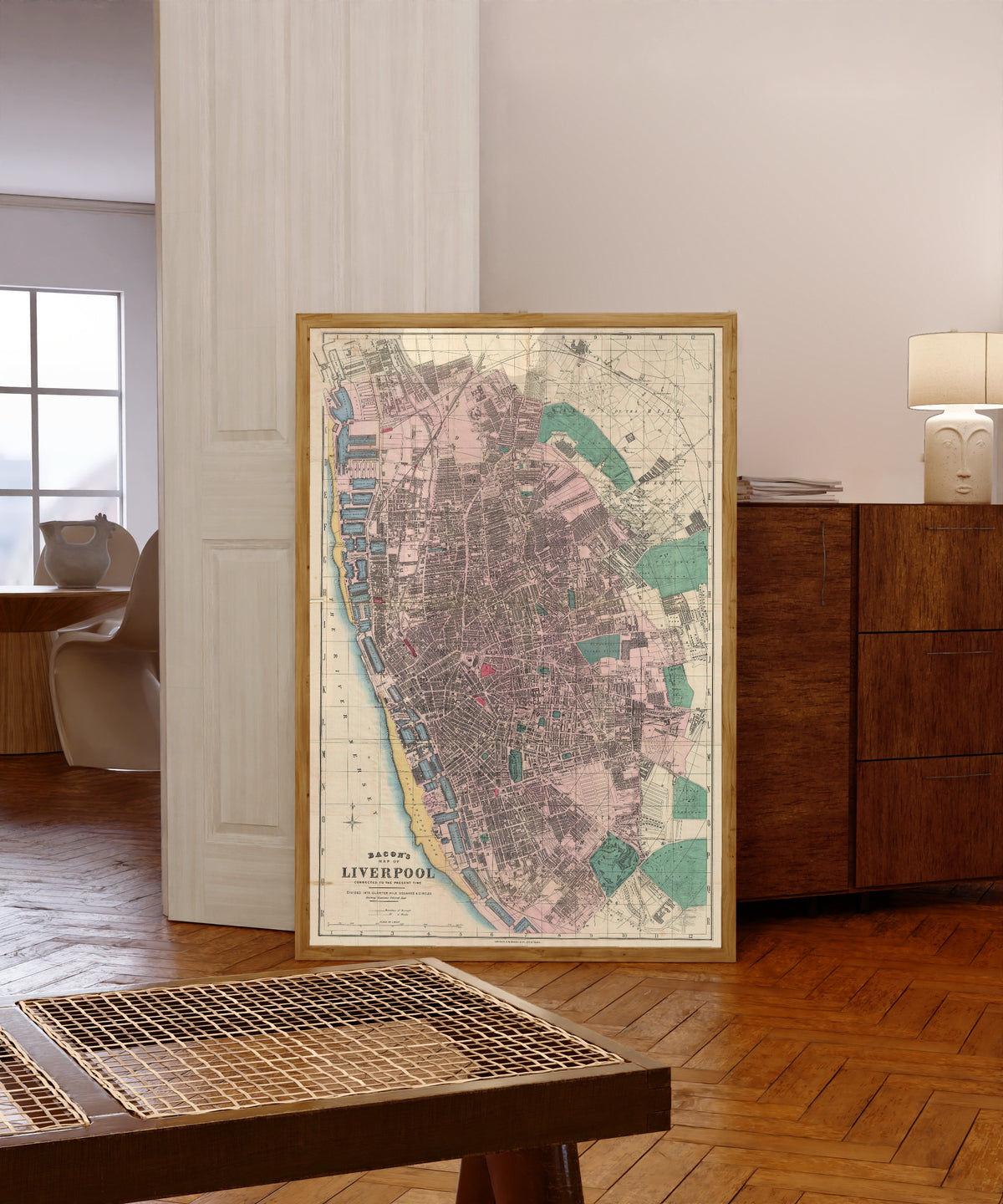 Framed art print of Bacon's Map of Liverpool by George Washington Bacon, in a light wood frame leaning in a warm room with a parquet floor