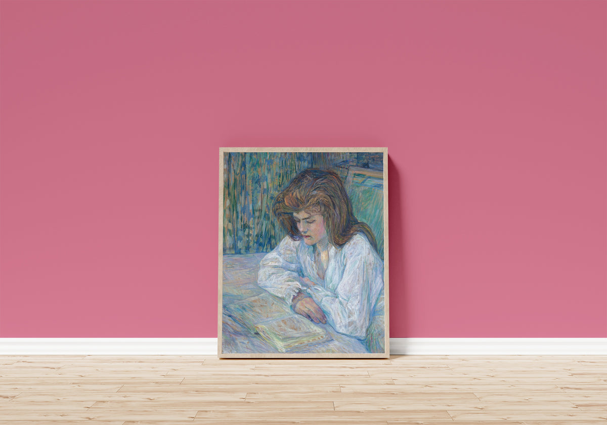 Framed art print of La Liseuse by Henri de Toulouse-Lautrec, in a light wood frame leaning on a pink wall above a wooden floor