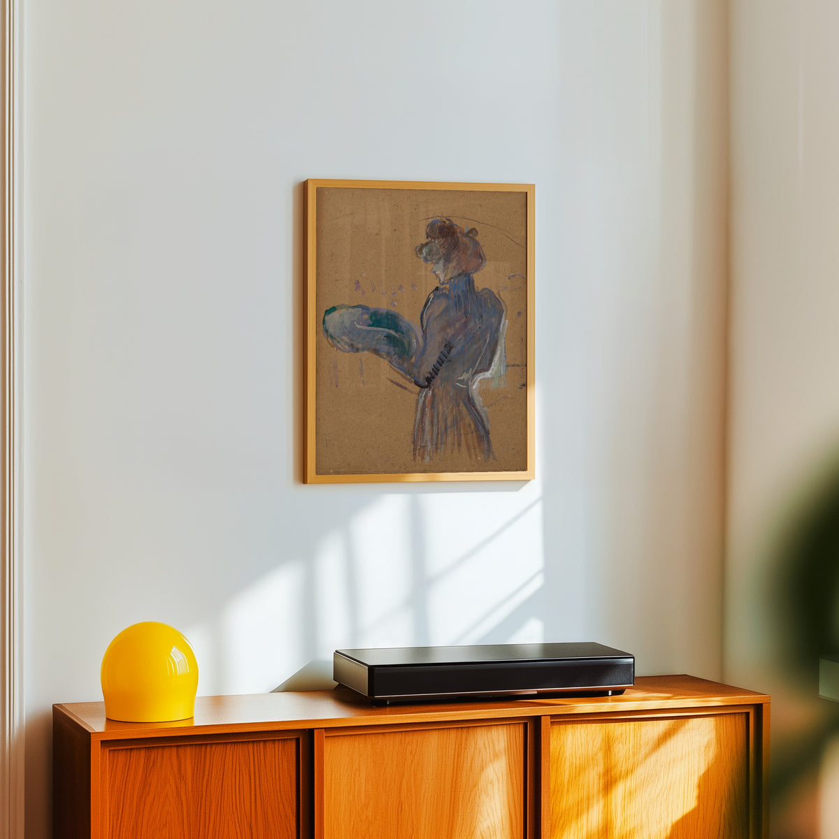 Framed art print of La Modiste, Mademoiselle Margouin by Henri de Toulouse-Lautrec, in a light wood frame on a white wall above an orange sideboard