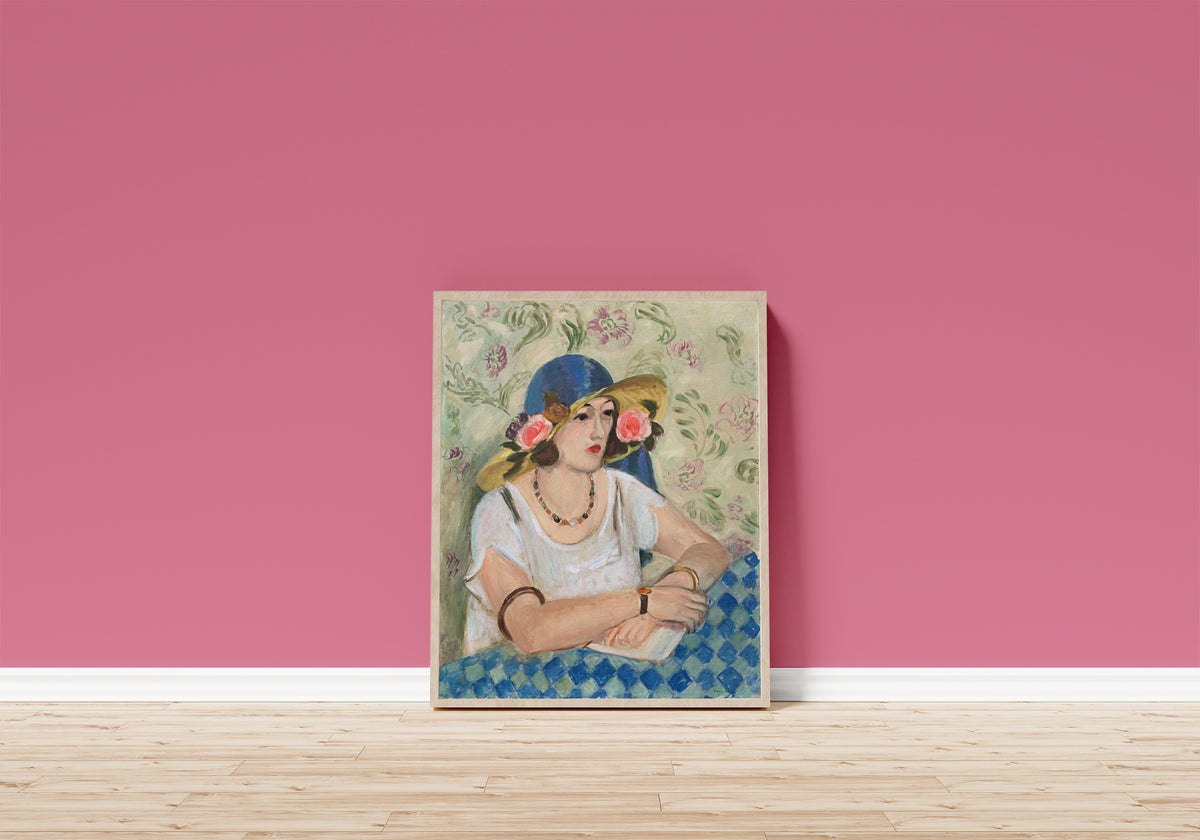 Framed art print of Woman with a Flowered Hat by Henri Matisse, in a light wood frame leaning on a pink wall above a wooden floor