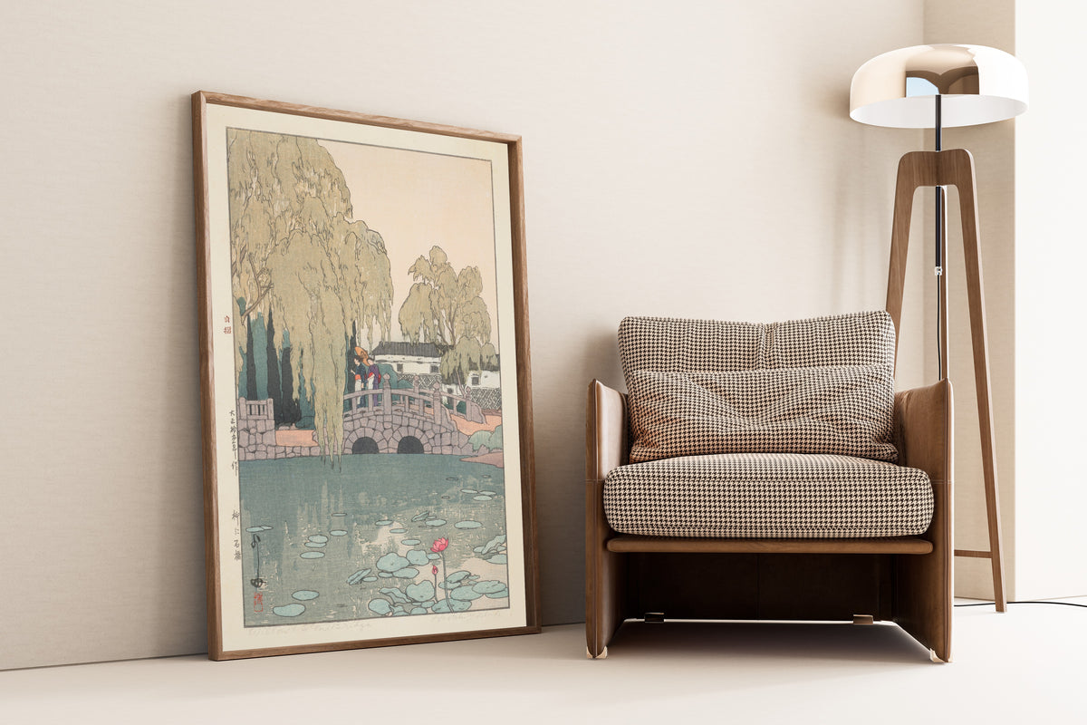Framed art print of Willow and Stone Bridge by Hiroshi Yoshida, leaning against a wall beside an armchair and a floor lamp