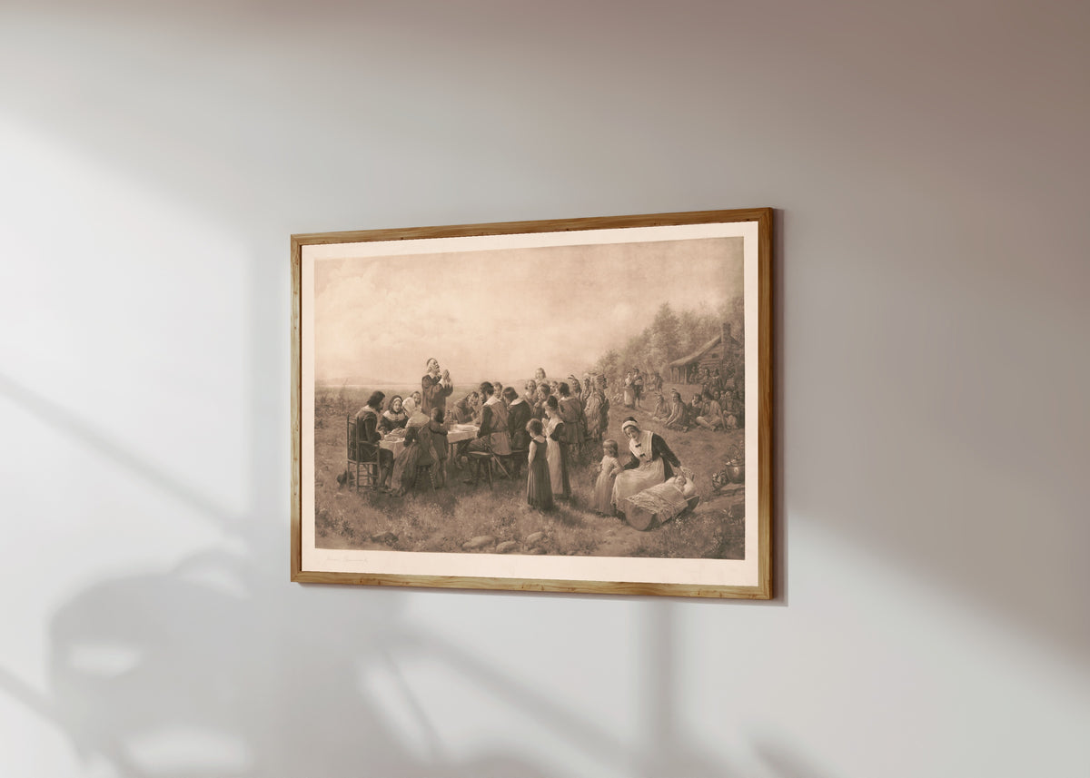 Framed art print of The First American Thanksgiving by Jennie Brownscombe, in a light wood frame on a sunlit white wall, seen at a slight angle