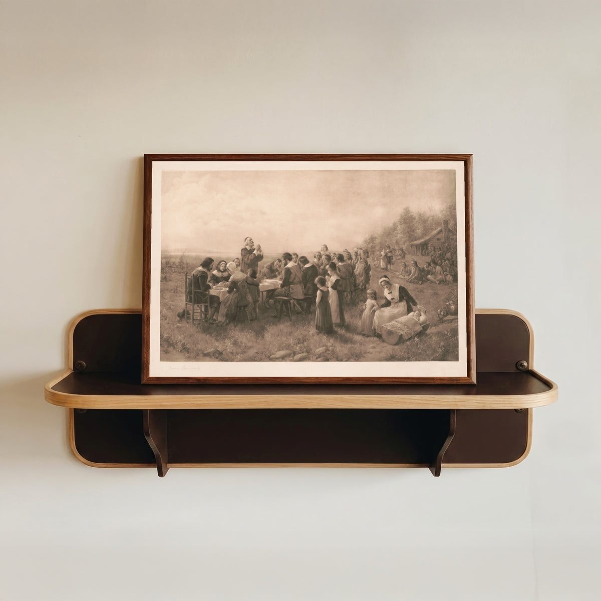 Framed art print of The First American Thanksgiving by Jennie Brownscombe, in a dark wood frame resting on a wall shelf