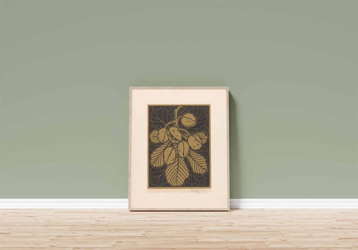 Framed art print of Hazelnuts and leaves by Julie de Graag, in a light wood frame leaning on a sage green wall above a wooden floor