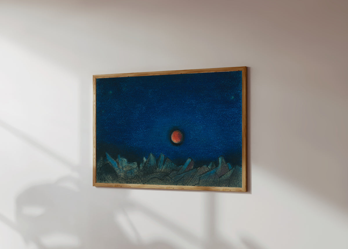 Framed art print of Untitled (Red Moon over a Landscape of Ruins) by Karl Wiener, in a light wood frame on a sunlit white wall, seen at a slight angle