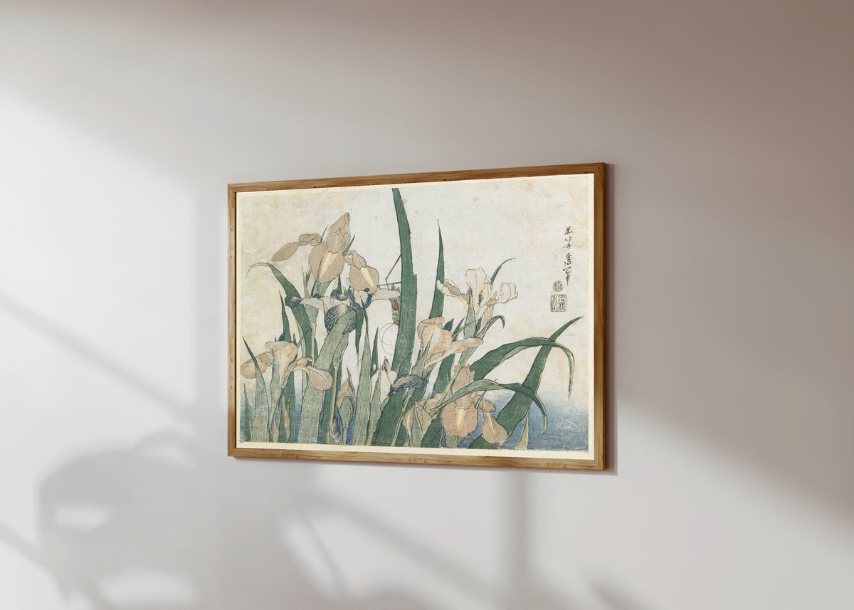 Framed art print of Large Flowers: Irises and Grasshopper by Katsushika Hokusai, in a light wood frame on a sunlit white wall, seen at a slight angle