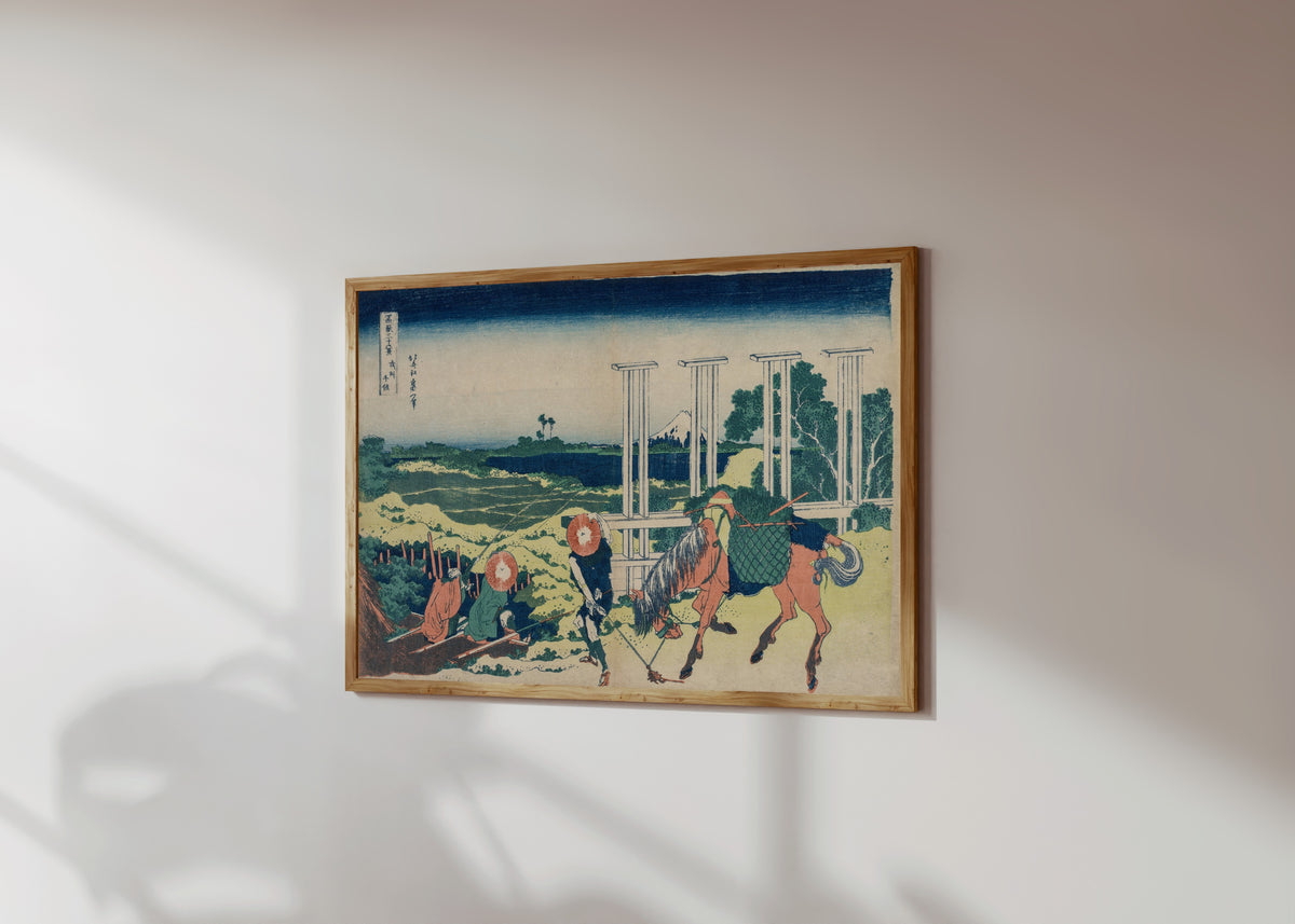 Framed art print of Thirty-six Views of Mount Fuji: The Weir at… by Katsushika Hokusai, in a light wood frame on a sunlit white wall, seen at a slight angle