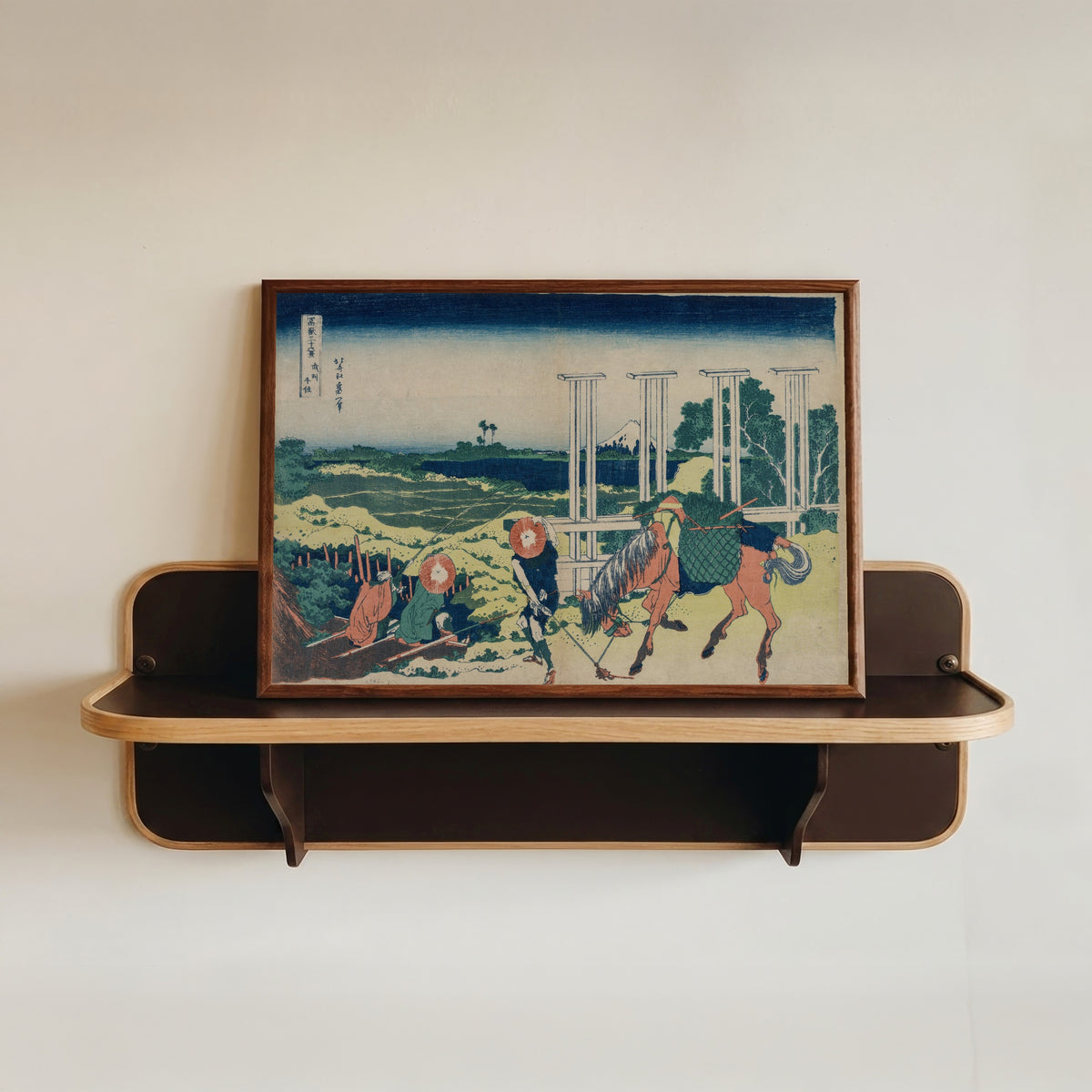Framed art print of Thirty-six Views of Mount Fuji: The Weir at Senju in Musashi Province by Katsushika Hokusai, in a dark wood frame resting on a wall shelf