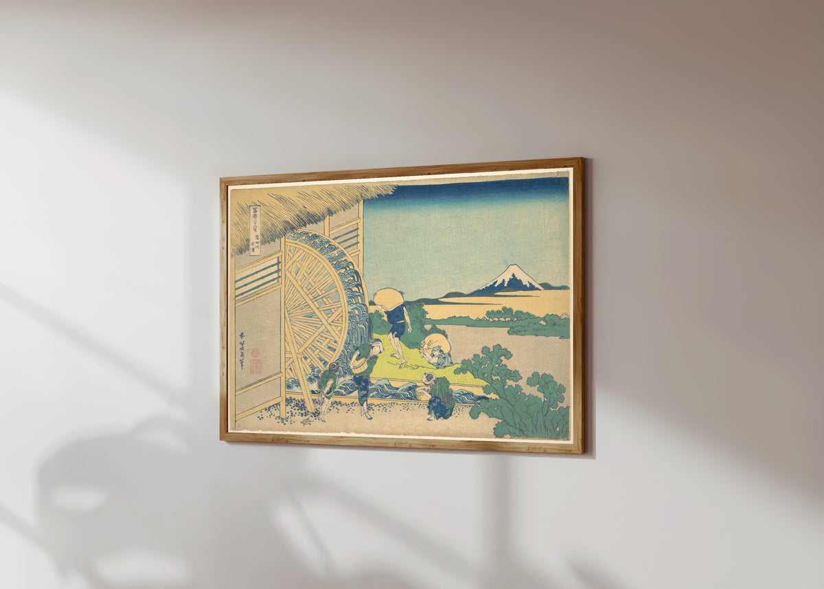 Framed art print of Thirty-six Views of Mount Fuji: Waterwheel at… by Katsushika Hokusai, in a light wood frame on a sunlit white wall, seen at a slight angle