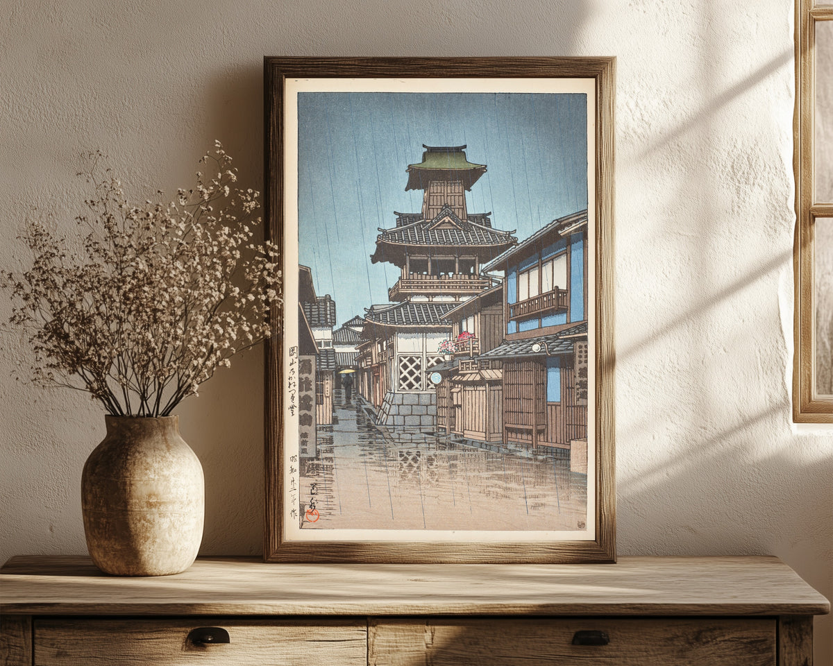 Framed art print of Bell Tower at Okayama by Kawase Hasui, in a dark wood frame leaning on a rustic console beside dried flowers