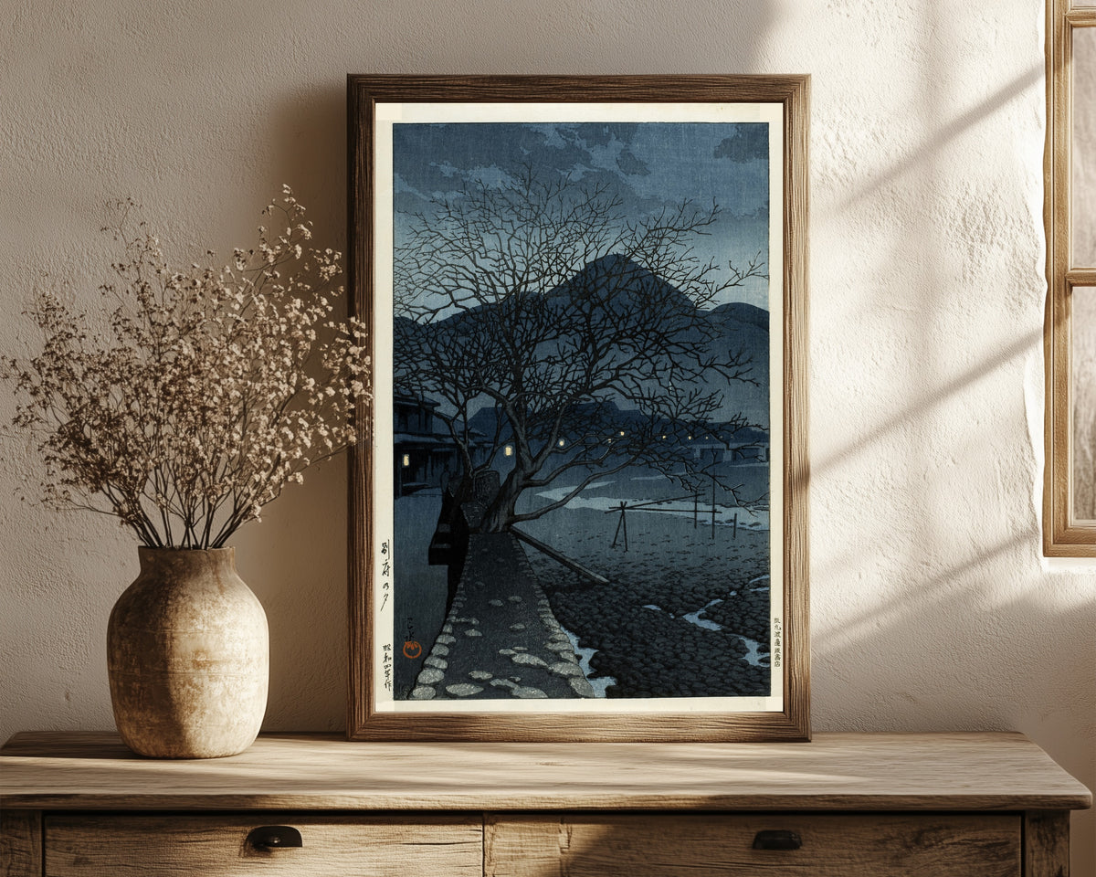 Framed art print of Evening in Beppu by Kawase Hasui, in a dark wood frame leaning on a rustic console beside dried flowers