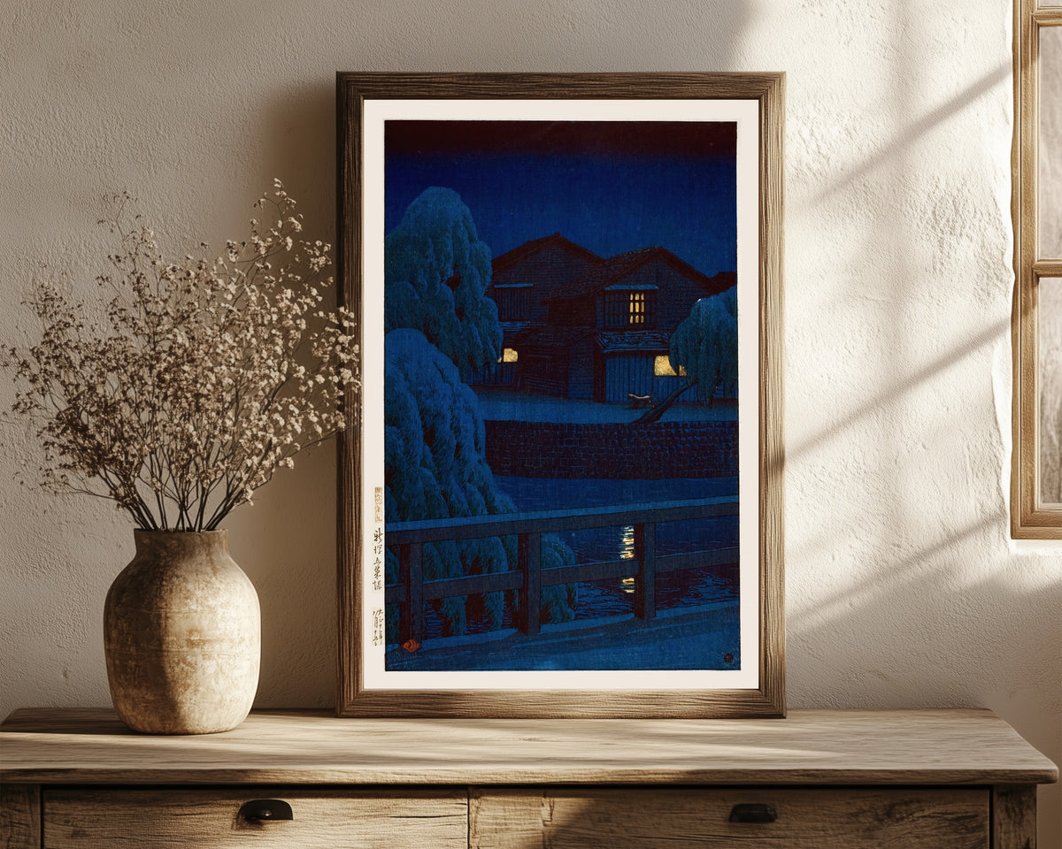 Framed art print of Gosai Canal, Niigata by Kawase Hasui, in a dark wood frame leaning on a rustic console beside dried flowers