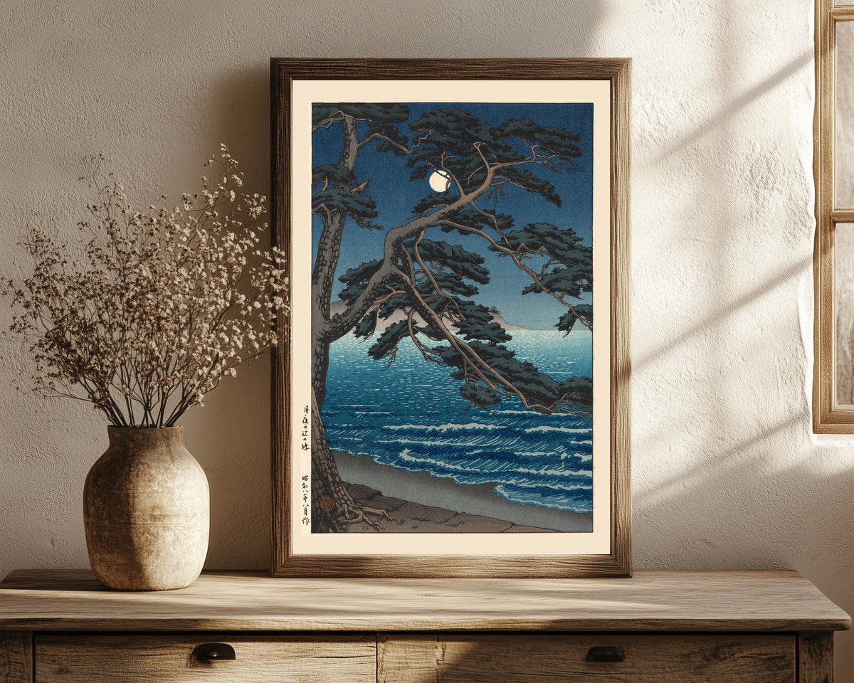 Framed art print of Moon at Enoshima Beach by Kawase Hasui, in a dark wood frame leaning on a rustic console beside dried flowers