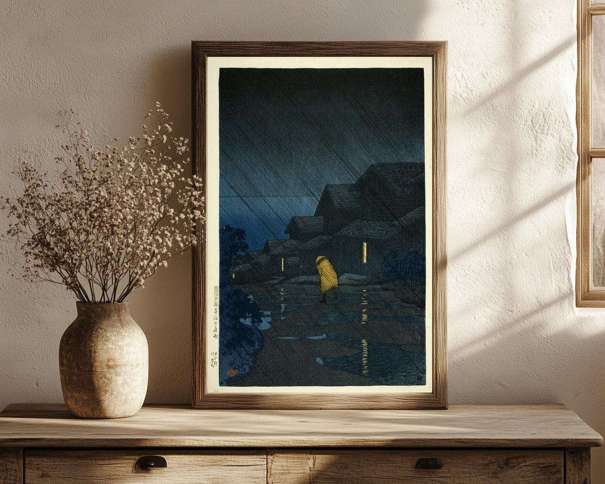 Framed art print of Rain at Teradomari Giclée Print: by Kawase Hasui, in a dark wood frame leaning on a rustic console beside dried flowers