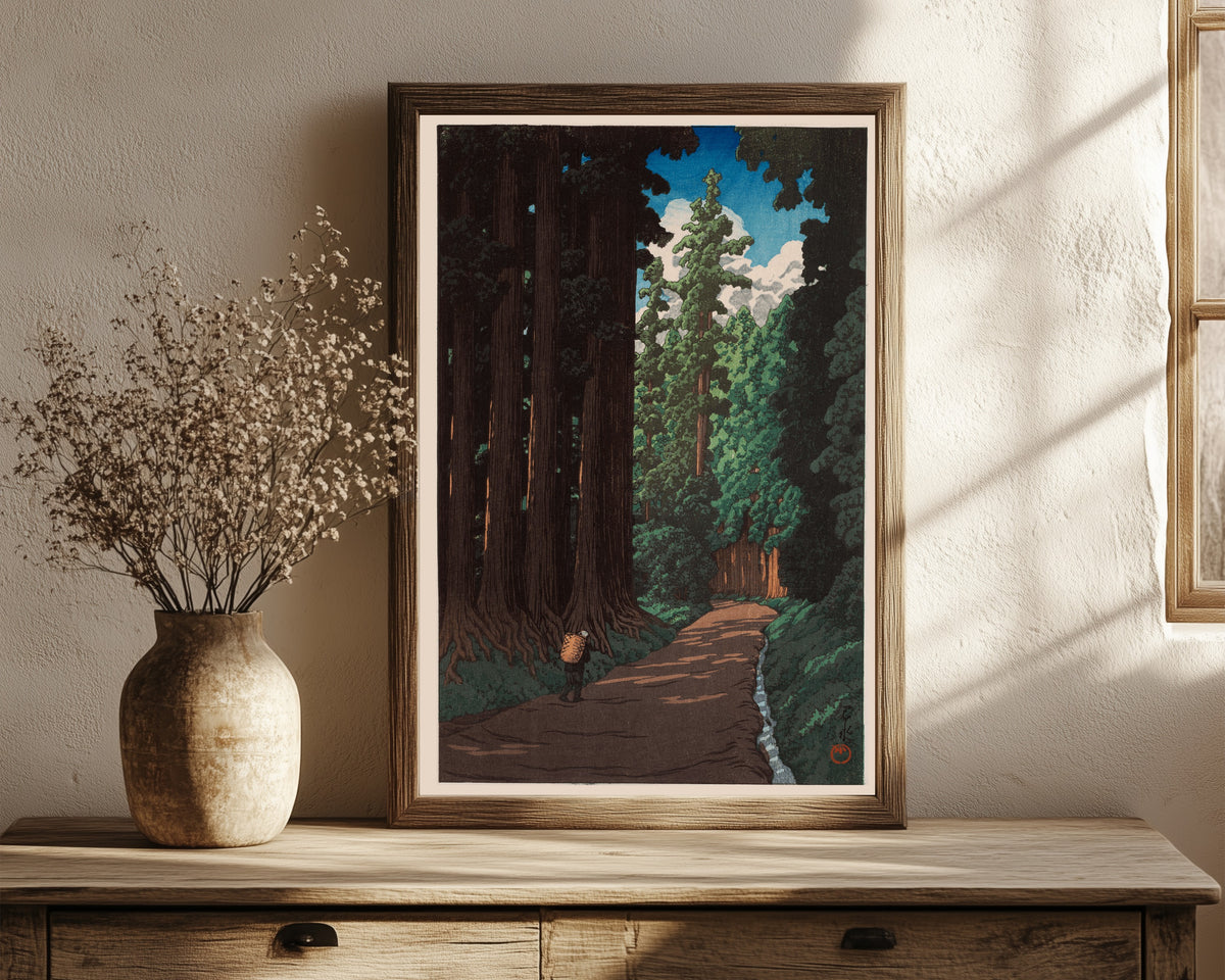 Framed art print of Road to Nikko by Kawase Hasui, in a dark wood frame leaning on a rustic console beside dried flowers