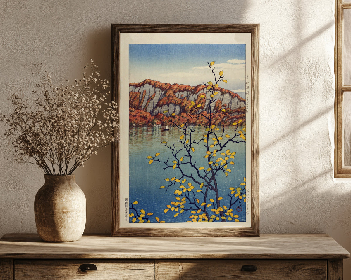 Framed art print of Lake Towada by Kawase Hasui, in a dark wood frame leaning on a rustic console beside dried flowers