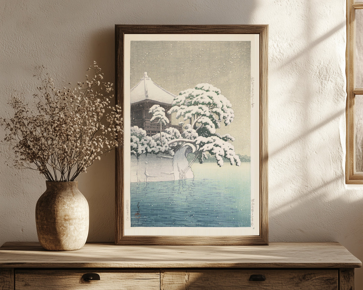 Framed art print of Snow at Godōidō, Matsushima by Kawase Hasui, in a dark wood frame leaning on a rustic console beside dried flowers