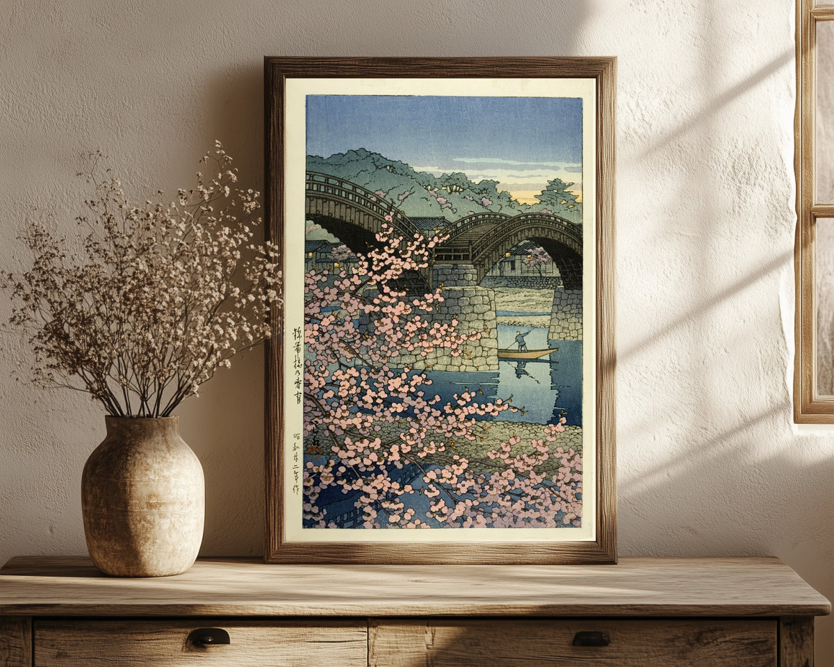 Framed art print of Kintai Bridge Cherry Blossoms by Kawase Hasui, in a dark wood frame leaning on a rustic console beside dried flowers
