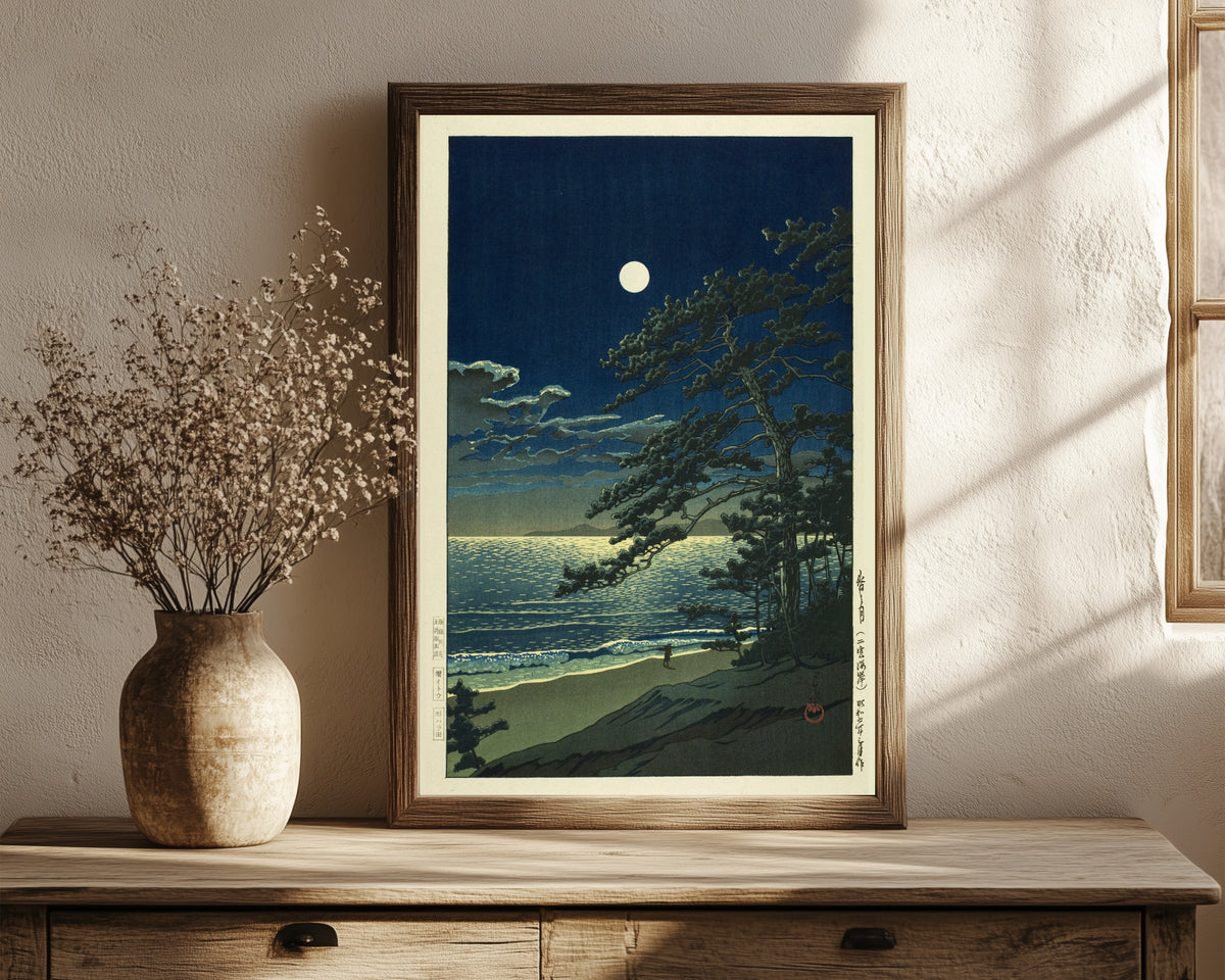 Framed art print of Spring Moon in Ninomiya Beach by Kawase Hasui, in a dark wood frame leaning on a rustic console beside dried flowers