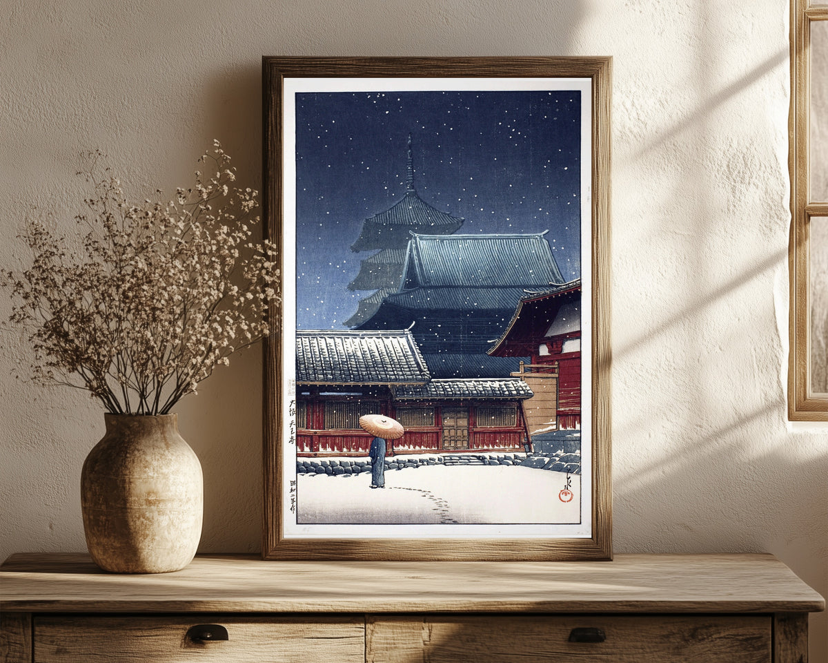 Framed art print of Tennoji Temple, Osaka, in Snow by Kawase Hasui, in a dark wood frame leaning on a rustic console beside dried flowers