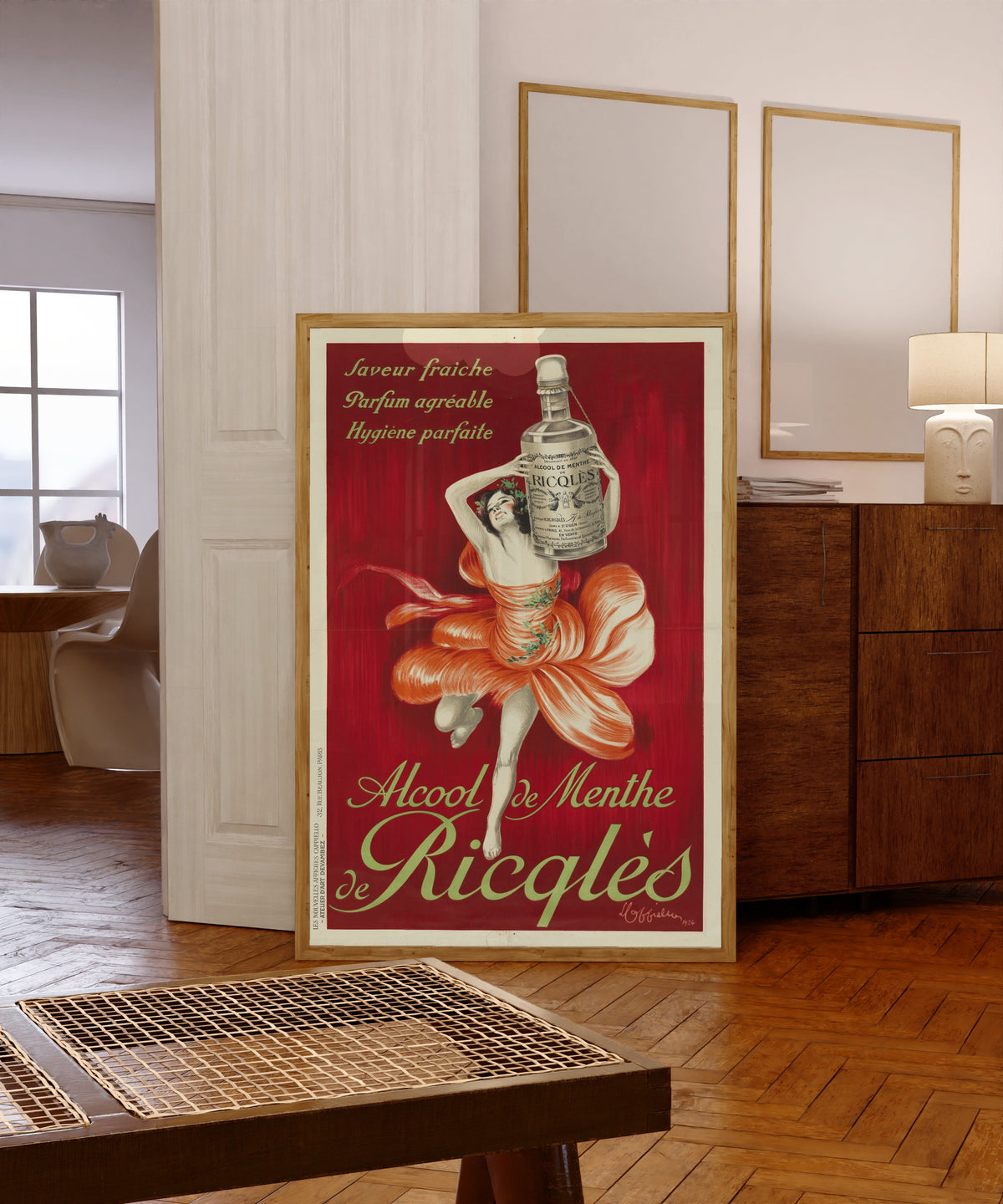 Framed art print of Alcool de menthe de Ricqlès by Leonetto Cappiello, in a light wood frame leaning in a warm room with a parquet floor