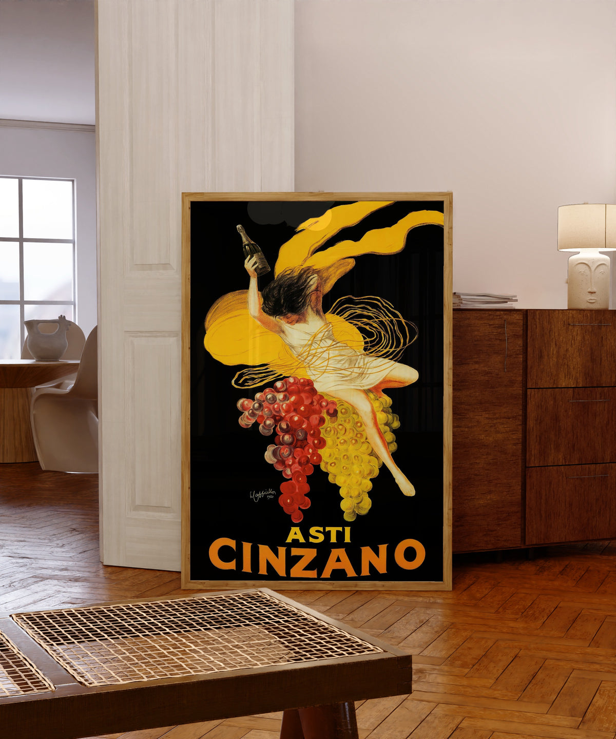 Framed art print of Asti Cinzano by Leonetto Cappiello, in a light wood frame leaning in a warm room with a parquet floor